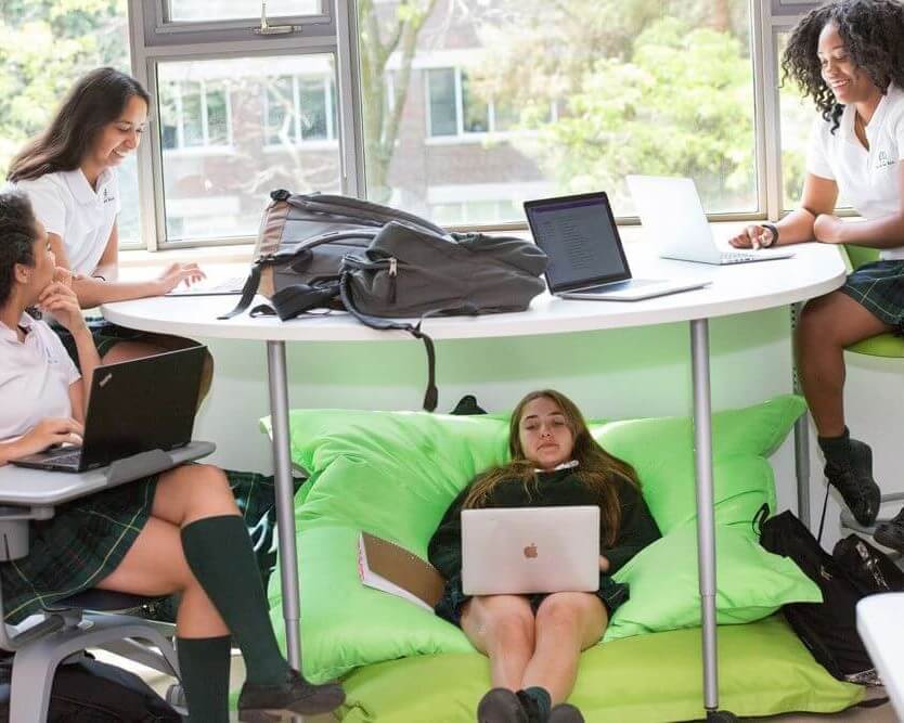 Four students in school uniforms work on laptops around a circular table by a window; one girl reclines on green bean bags under the table, using her laptop, while the others sit on chairs above, smiling and chatting.