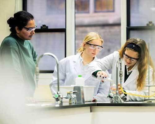 Three women wearing safety goggles work at a laboratory bench; one observes while the other two, in lab coats, focus on conducting an experiment using lab equipment and a liquid.
