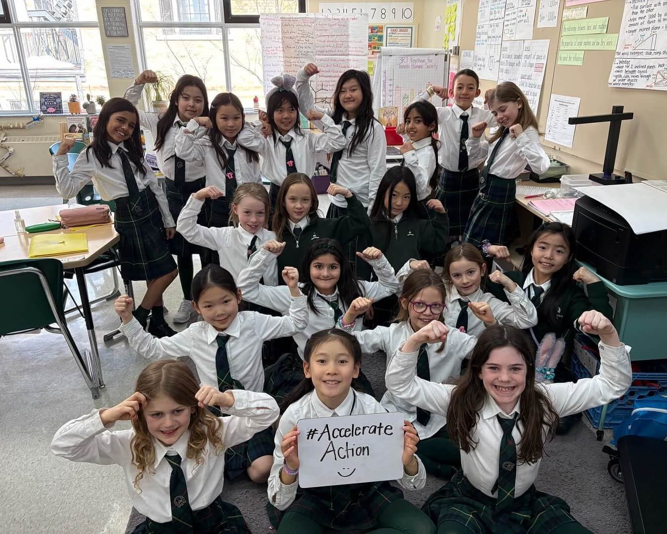 A group of schoolgirls in uniforms pose in a classroom, smiling and flexing their arms. One girl in front holds a sign reading "#Accelerate Action" with a smiley face. Posters and papers decorate the classroom walls.