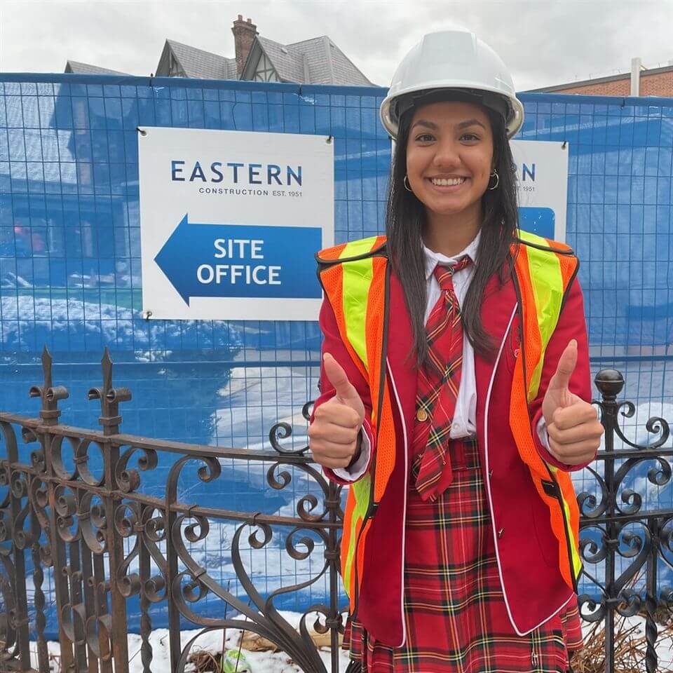 A smiling young woman in a red plaid uniform, orange safety vest, and white hard hat gives two thumbs up in front of a construction site and a sign reading "Eastern Construction Site Office.