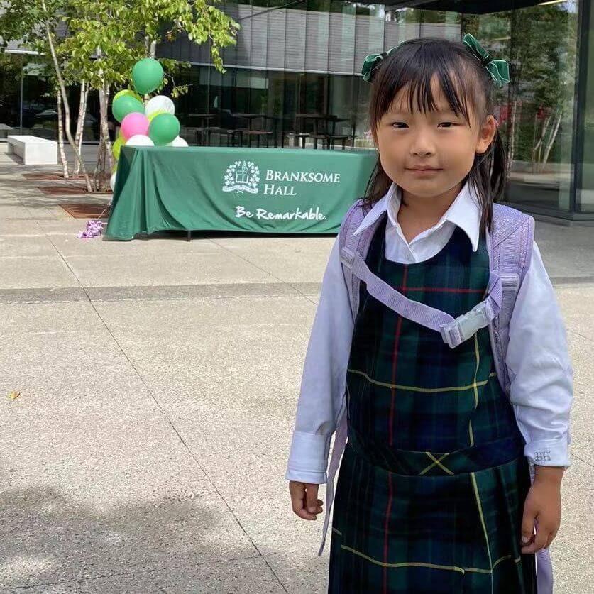 A young girl in a green plaid school uniform and purple backpack stands outdoors, smiling. Behind her is a table with balloons and a green cloth that reads "Branksome Hall. Be Remarkable." Trees are in the background.