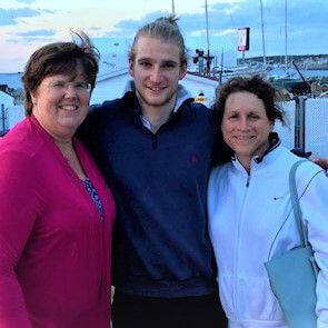 Three people stand close together outdoors, smiling at the camera. The man in the center has long blond hair, while the women on either side wear glasses and casual jackets. A marina with boats and a cloudy sky is in the background.