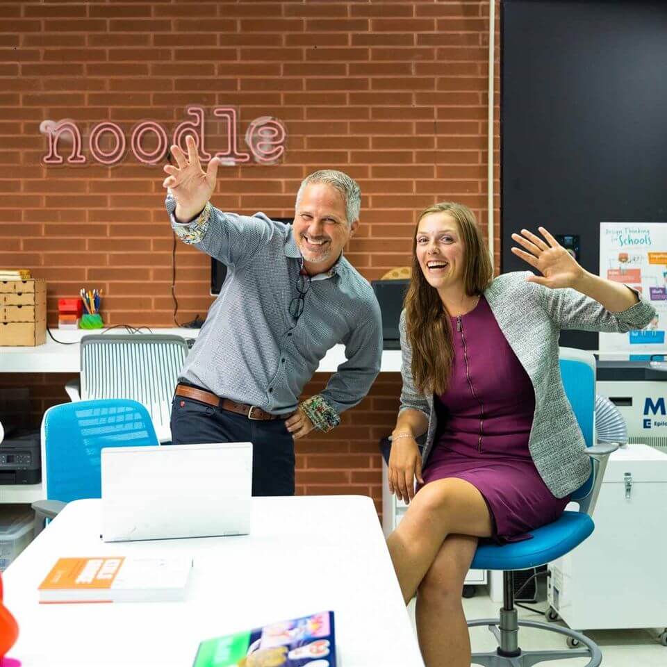 Two smiling adults, one standing and one sitting on a chair, wave enthusiastically in a bright office space with a brick wall and a neon "noodle" sign in the background.