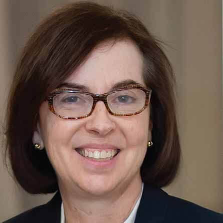 A woman with short brown hair, wearing tortoiseshell glasses, small round earrings, and a dark blazer, smiles at the camera against a neutral background.