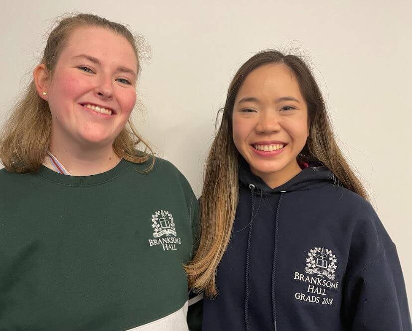 Two young women smiling, wearing Branksome Hall sweatshirts; one in a dark green top, the other in a navy hoodie that says "BRANKSOME HALL GRADS 2018." They stand against a plain, light-colored background.