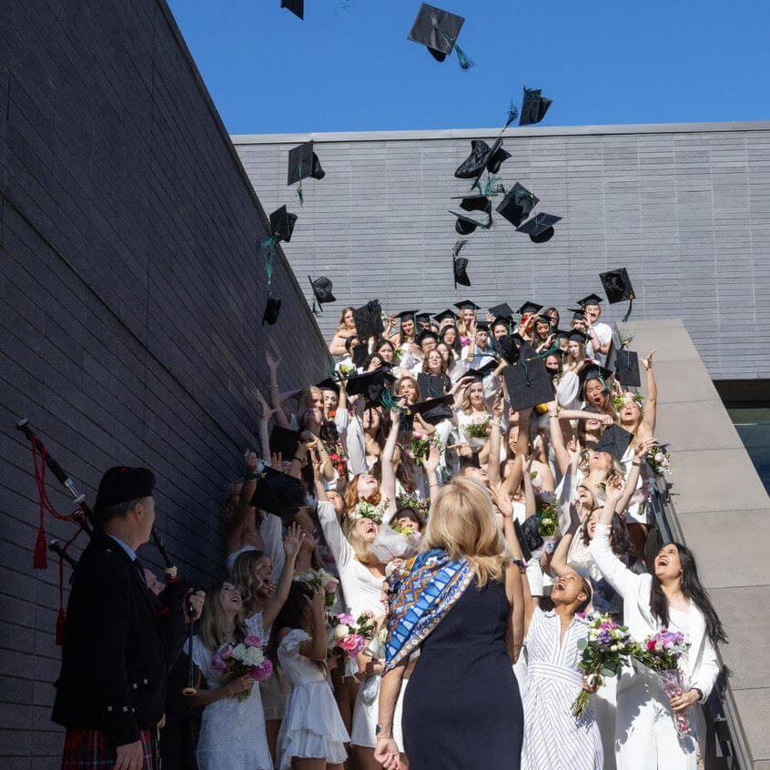 A group of graduates in white attire joyfully toss their caps into the air on outdoor steps, holding bouquets and celebrating, while a person in a kilt plays bagpipes nearby under a clear blue sky.