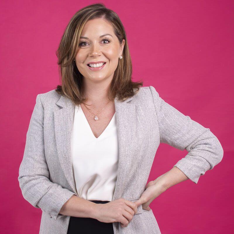 A smiling woman with shoulder-length brown hair stands in front of a bright pink background, wearing a light gray blazer over a white blouse with one hand on her hip.