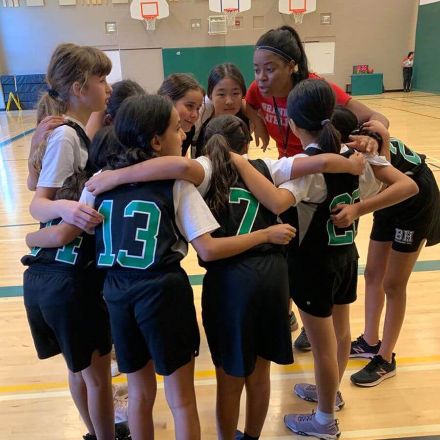 A coach in a red shirt talks to a group of young girls in basketball uniforms standing in a huddle on an indoor basketball court, preparing for a game or practice.