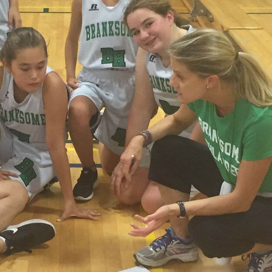 A basketball coach kneels on a gym floor, talking to young players in Branksome Hall uniforms during a timeout. The players listen attentively, seated and kneeling around the coach.