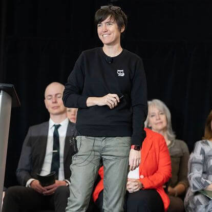 A person in casual clothing stands and smiles on stage, holding a small device. Several people are seated behind them, including a man in a suit and women in business attire, against a dark background.