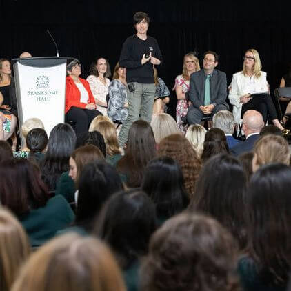 A person stands and speaks with a microphone on stage in front of a seated panel and a large audience at Branksome Hall.