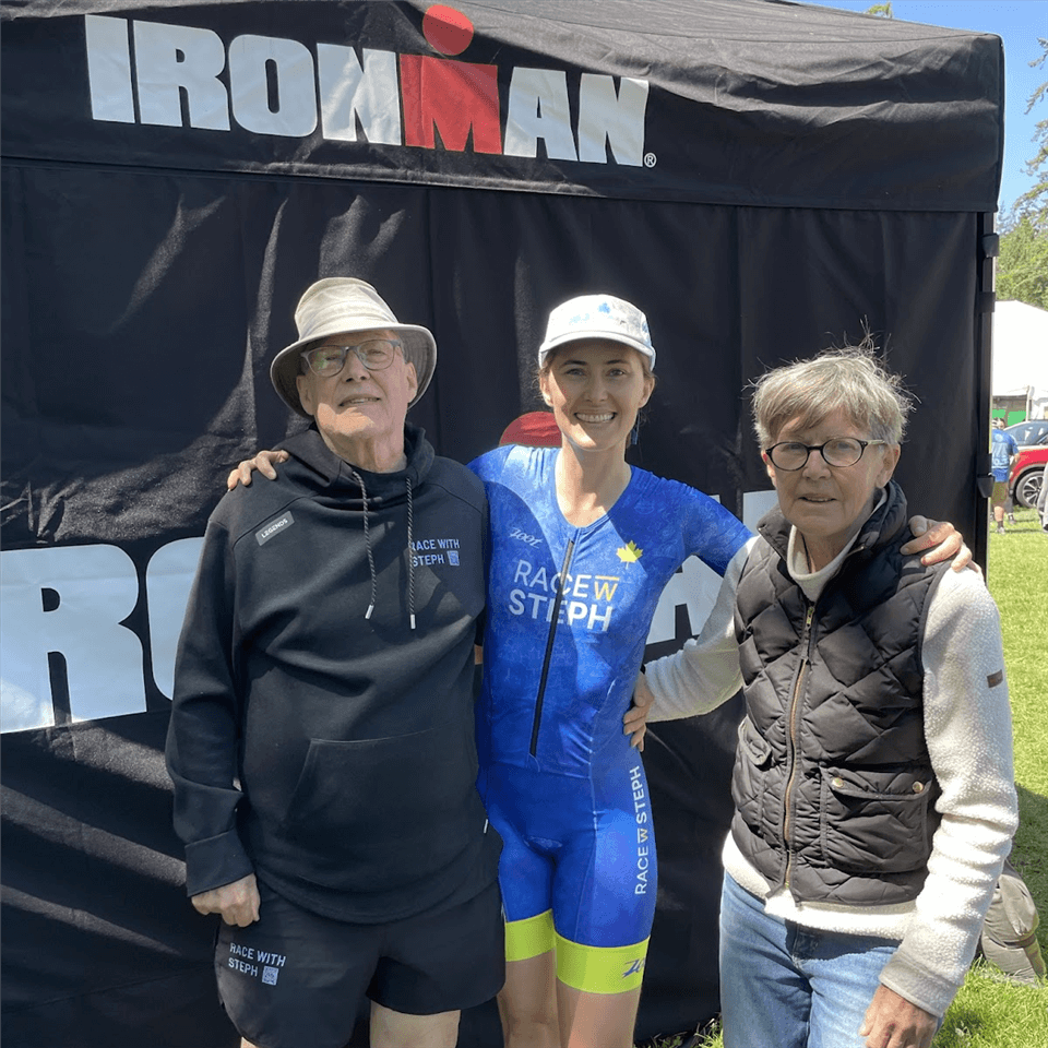 Three people stand smiling in front of a tent with an IRONMAN logo. The person in the middle is wearing a blue athletic outfit and a cap, while the two older people on either side wear glasses and casual outdoor clothing.