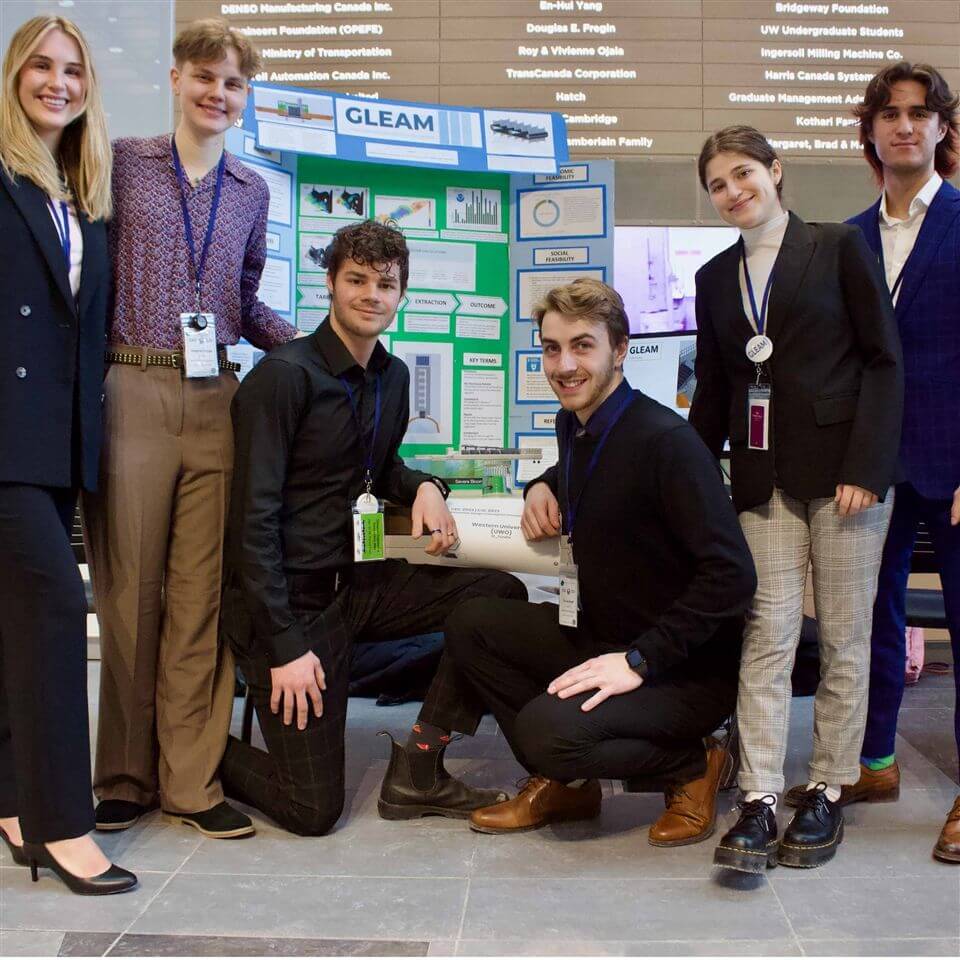 Six young adults, dressed in business attire, pose and smile around a science fair project display board titled “GLEAM” in a well-lit indoor space with a wall of donor names behind them.