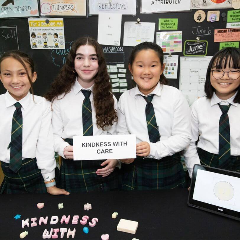 Four smiling students in school uniforms stand behind a table with "KINDNESS WITH CARE" sign and decorated letters. Classroom walls display colorful posters and notes in the background.