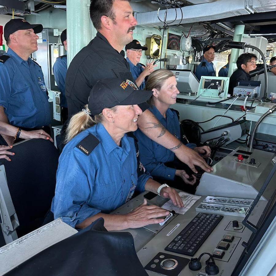 A group of navy personnel in blue uniforms work together at control panels inside a ship’s bridge, focusing on screens and controls, with some smiling and collaborating.