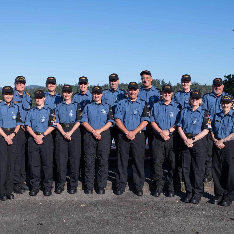 A group of fifteen people in matching blue shirts, dark pants, and caps stand together outdoors, smiling at the camera with a clear blue sky in the background.