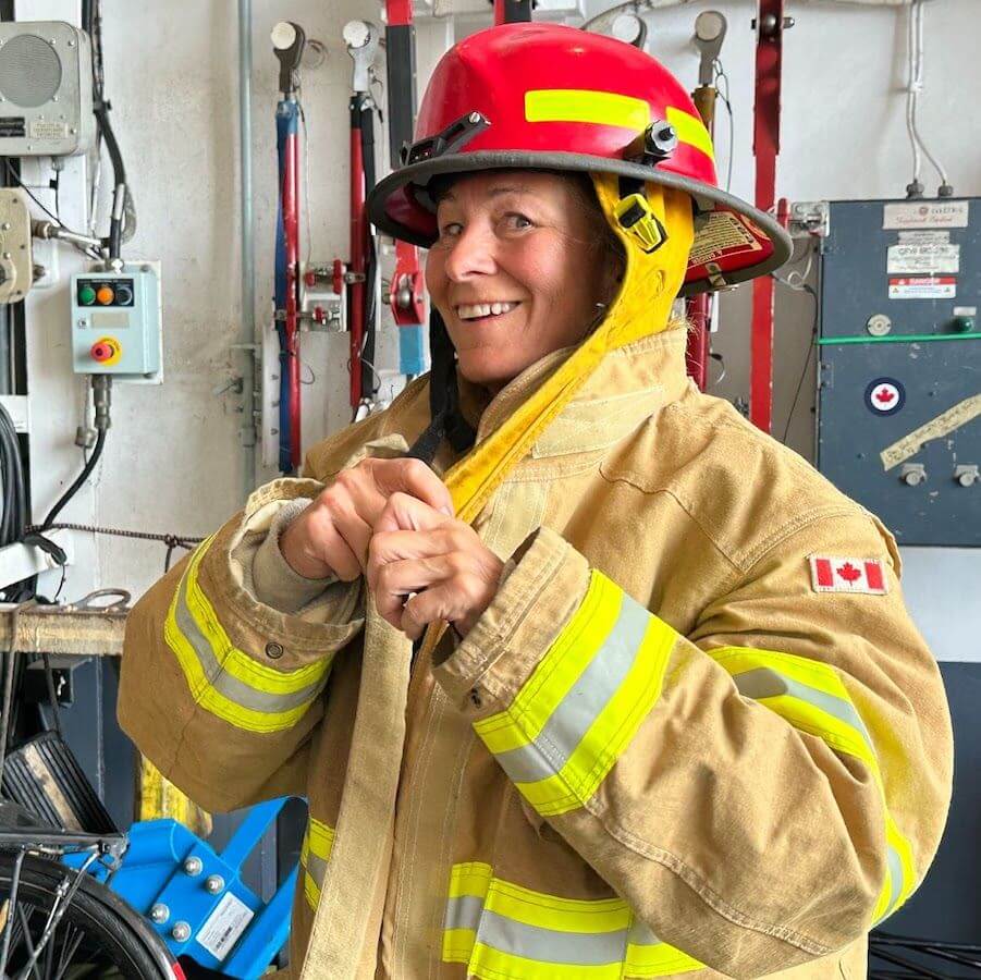 A smiling woman in a firefighter’s uniform and red helmet adjusts her jacket inside a fire station. She stands near equipment and wears a jacket with a Canadian flag patch on the sleeve.