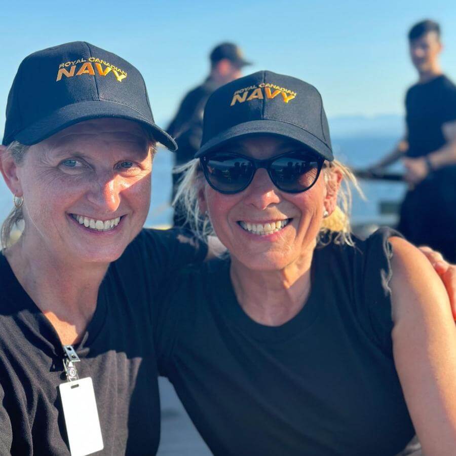 Two smiling women wearing black "Royal Canadian Navy" hats and black shirts sit close together outdoors on a sunny day. Other people in similar attire are visible in the background.