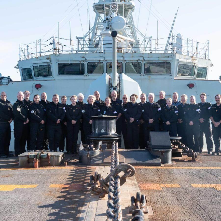 A large group of uniformed crew members stands together on the deck of a ship, posing for a photo in front of the ship’s bridge and anchor chain, under clear daylight.