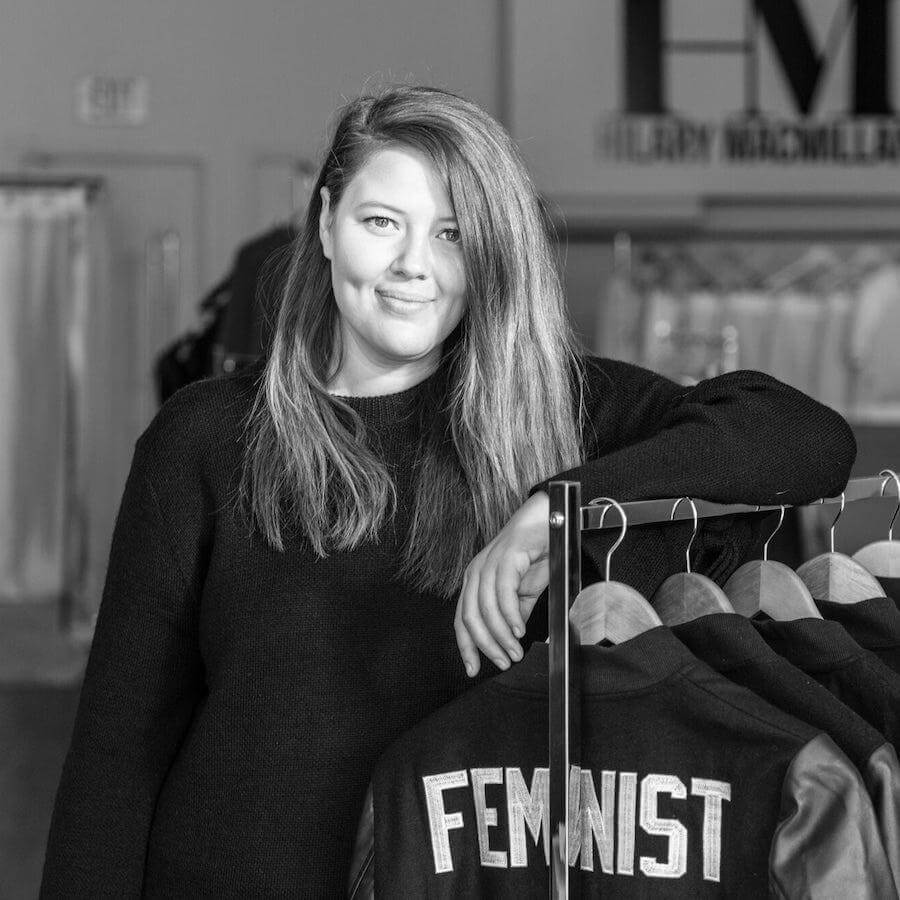 A woman with long hair, wearing a black sweater, stands next to a clothing rack with jackets that have "FEMINIST" written on the back. The image is in black and white.