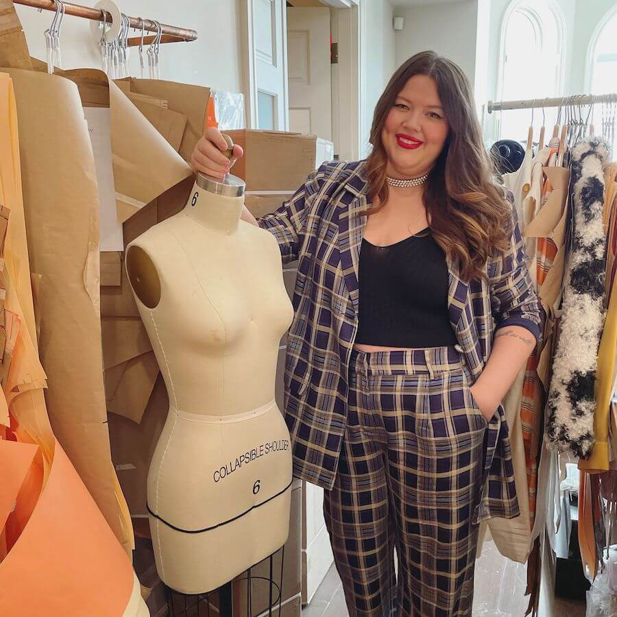 A woman with long brown hair, wearing a plaid suit and black top, stands smiling with one hand on a dress form in a fashion studio filled with sewing patterns and clothes.