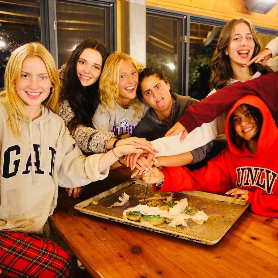 Six smiling young women gather around a wooden table, placing their hands together over a baking tray with remnants of a decorated cake, suggesting celebration and camaraderie.
