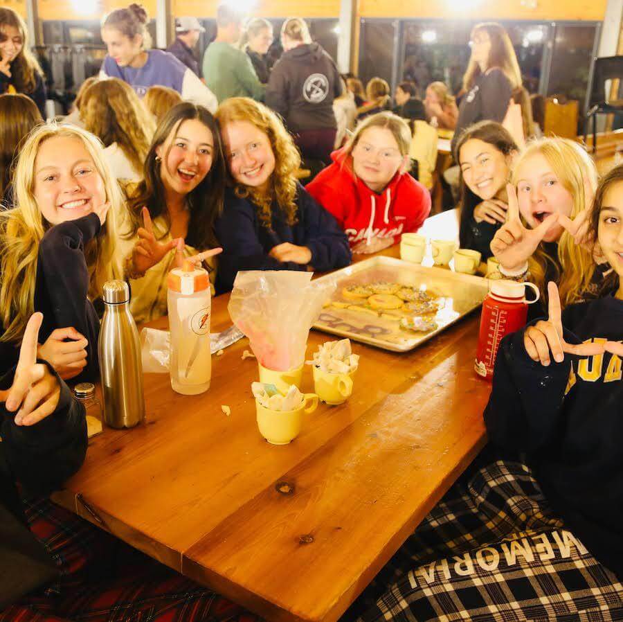 A group of smiling young people sit around a wooden table inside a bright, bustling room, with mugs, bottles, and a tray of cookies in front of them. Most are pointing upwards and appear to be enjoying themselves.