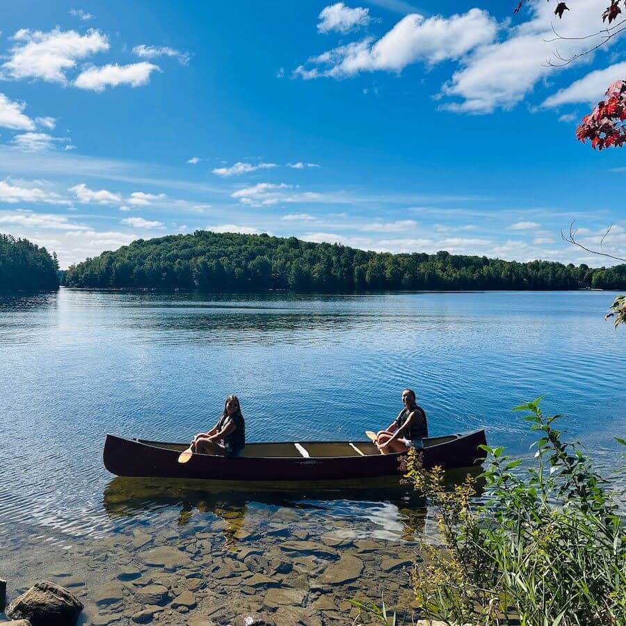 Two people sit in a canoe on a calm lake, surrounded by trees under a blue sky with scattered clouds. The shoreline with rocks and green foliage is visible in the foreground.