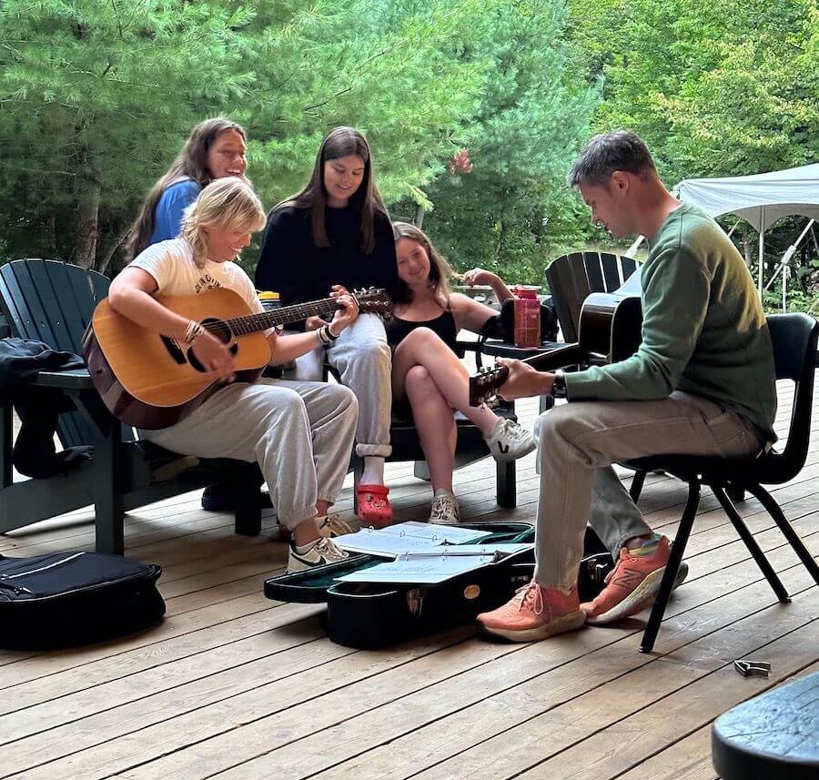 Four people sit outdoors on a wooden deck surrounded by trees. Three play acoustic guitars while one listens, smiling. Open guitar cases and sheet music are on the ground. The mood is relaxed and friendly.