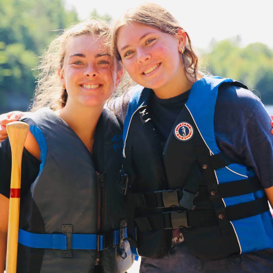 Two smiling young women wearing life jackets stand close together outdoors, with one holding a canoe paddle. Green trees and water are visible blurred in the background on a sunny day.