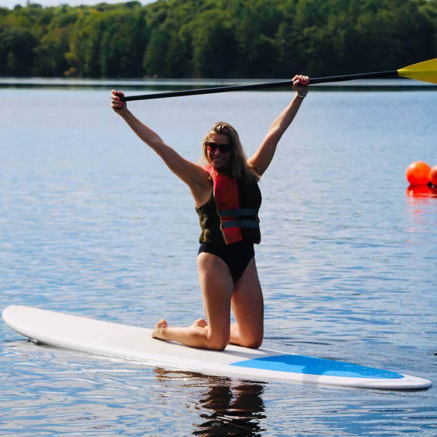 A woman kneels on a paddleboard in a lake, wearing a life vest and swimsuit, smiling with a raised paddle above her head. Trees and orange buoys are visible in the background.