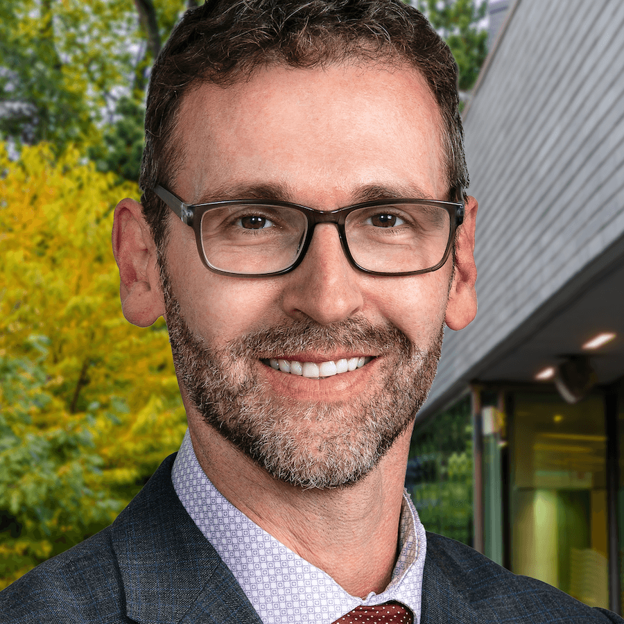 A smiling man with short curly brown hair, beard, and glasses, wearing a checked suit jacket, white dress shirt, and red tie, standing outdoors near a modern building with green trees in the background.
