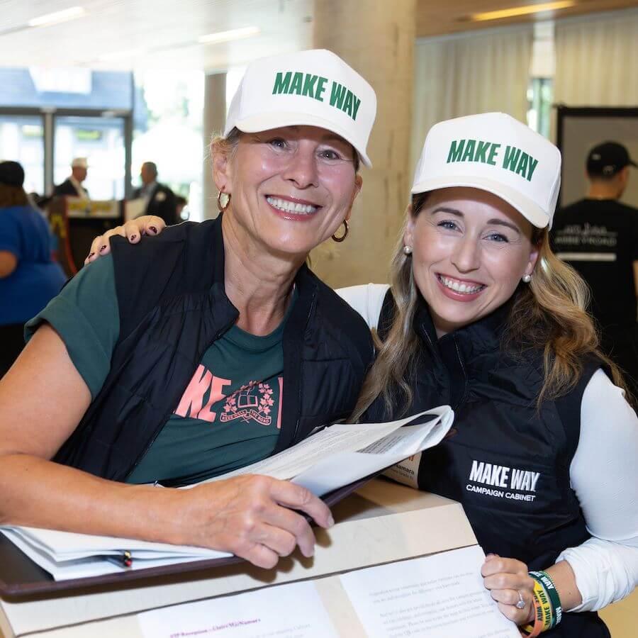 Two women wearing white "MAKE WAY" hats and black vests smile at the camera while holding a binder. They appear to be at an indoor event or registration desk, with people and tables in the background.