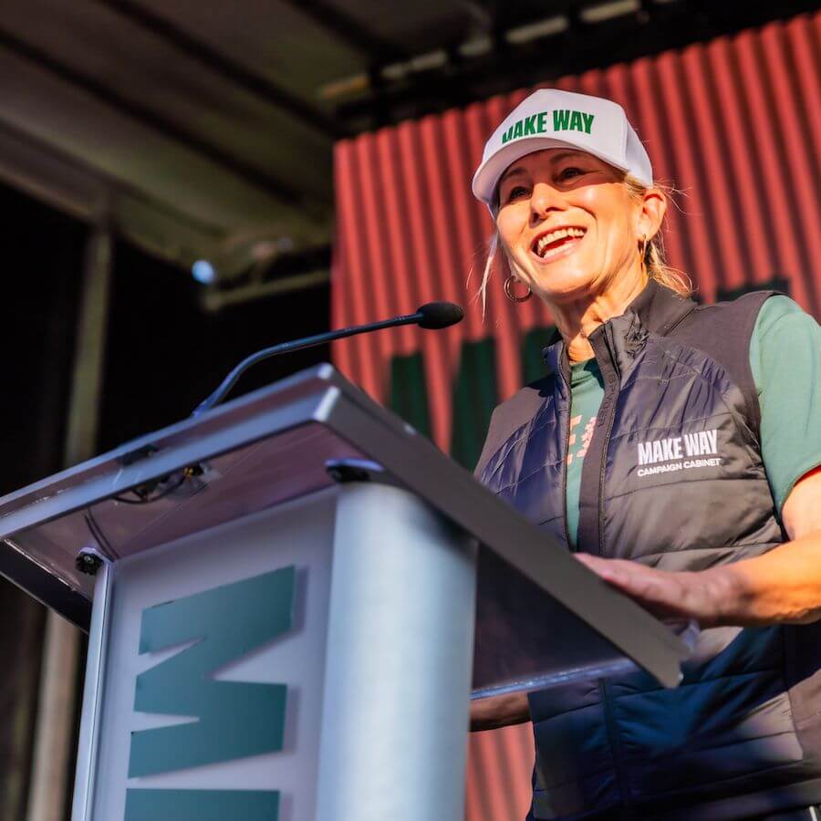 A smiling woman wearing a "MAKE WAY" hat and vest speaks at a podium during an outdoor event, with a red and green striped backdrop behind her.
