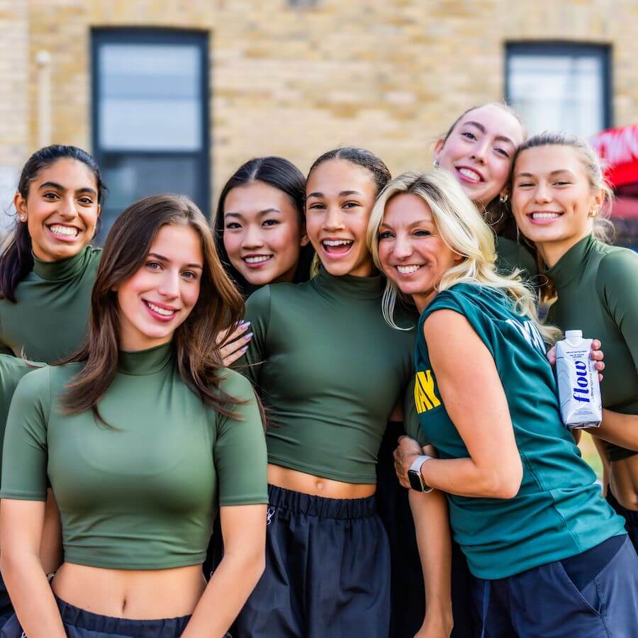 A group of six young women in matching green crop tops and one woman in a green T-shirt smile together outdoors, posing for a photo. One holds a drink. There’s a building and a red canopy in the background.