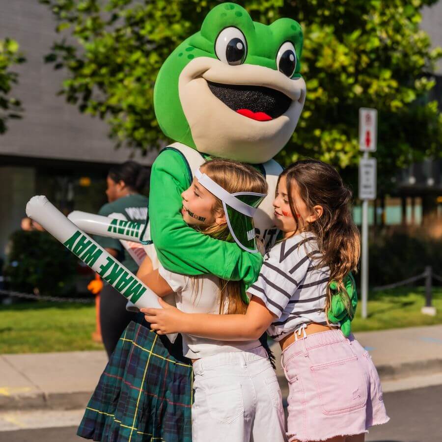 Two smiling girls hug a person in a large green frog mascot costume outdoors on a sunny day. One girl holds foam sticks that say “MAKE WAY.” Trees and a building appear in the background.