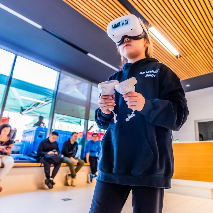 A child wearing a VR headset labeled "MAKE WAY" and holding controllers stands indoors, while several people sit in the background. Bright natural light enters through large windows.