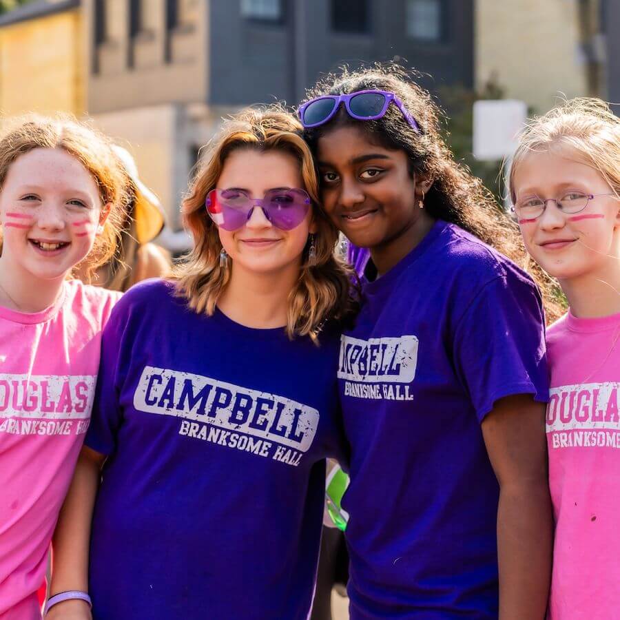 Four smiling girls pose outdoors. Two wear bright pink "DOUGLAS BRANKSOME HALL" shirts, and two wear purple "CAMPBELL BRANKSOME HALL" shirts. They stand close together, enjoying a sunny day.