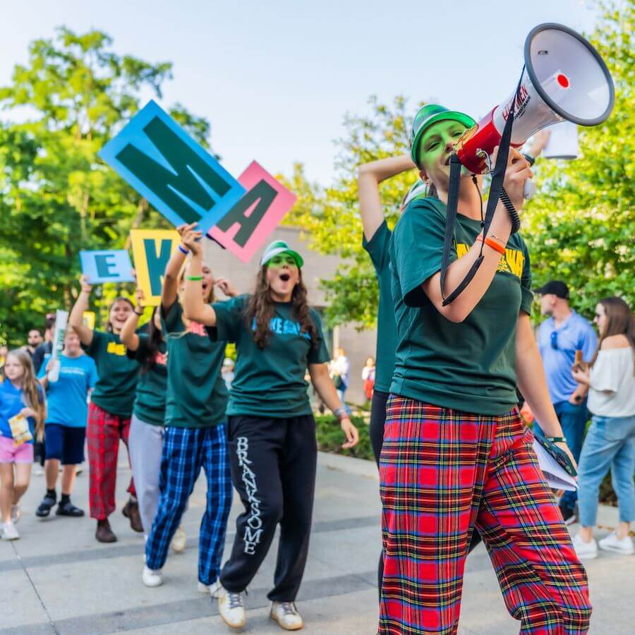 A group of people in colorful pajamas and green hats parade outdoors, holding large letter signs and a megaphone. Trees and a crowd are visible in the background on a sunny day.