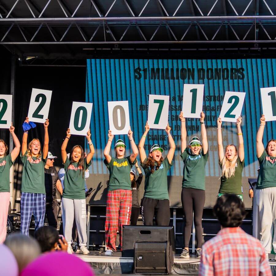 A group of people in green shirts and colorful pants hold up large signs with the numbers “22007121” onstage, celebrating in front of a large screen and cheering crowd at an outdoor event.