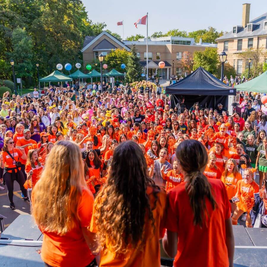 A large crowd of people, many wearing orange shirts, gathers outdoors in front of a stage with four people on it. Tents, trees, and buildings with Canadian flags are visible in the background.