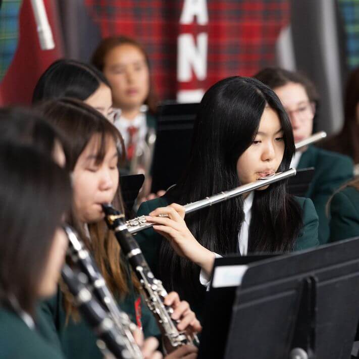 A group of students in green uniforms play musical instruments, including flutes and clarinets, during a band performance. A blurred red and black plaid banner is visible in the background.