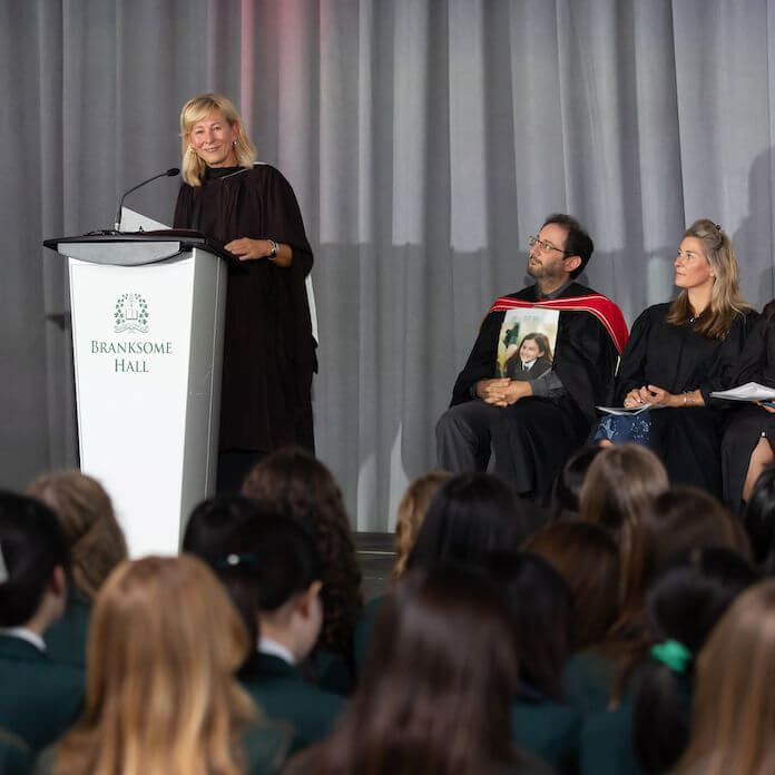 A woman speaks at a podium labeled "Branksome Hall" on a stage, while two seated adults and an audience of students in green uniforms listen attentively. A grey curtain hangs in the background.