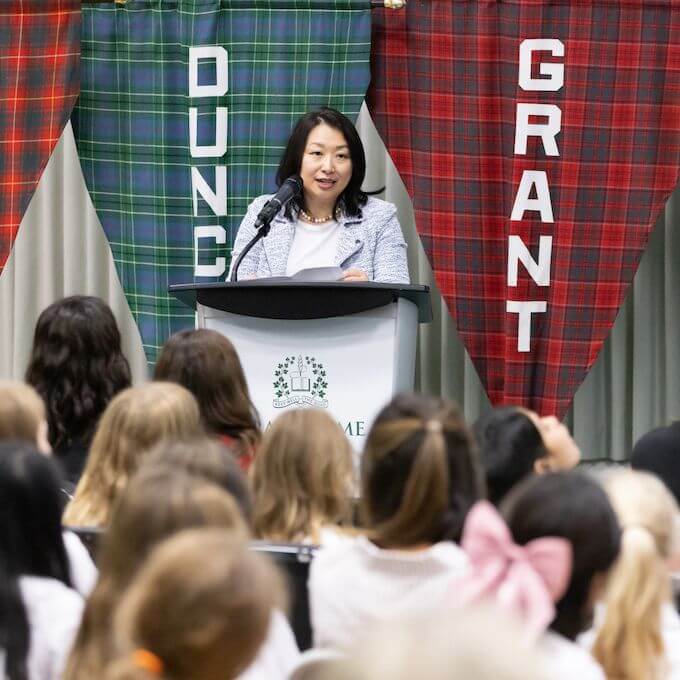 A woman speaks at a podium in front of an audience of children. Behind her hang two plaid banners reading "DUN" and "GRANT." The children face the speaker, some with bows in their hair.