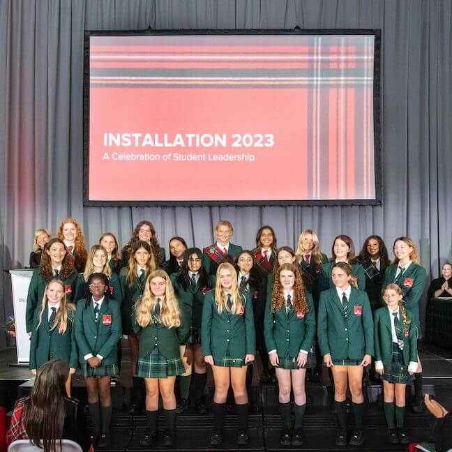 A group of students in green blazers pose together on stage in front of a screen that reads “INSTALLATION 2023: A Celebration of Student Leadership.” The group consists mostly of girls, all smiling.