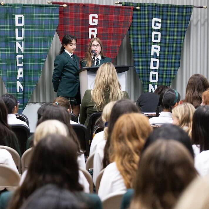 Two students in green blazers stand at a podium speaking to an audience. Behind them hang three plaid banners labeled "Duncan," "Grant," and "Gordon." The audience is seated, facing the speakers.