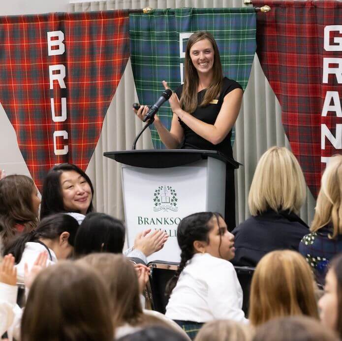 A woman stands at a podium labeled "Branksome Hall" smiling and clapping. She faces an audience of seated people, some clapping. Red and green plaid banners hang behind her, labeled "BRUCE" and "GRANT.