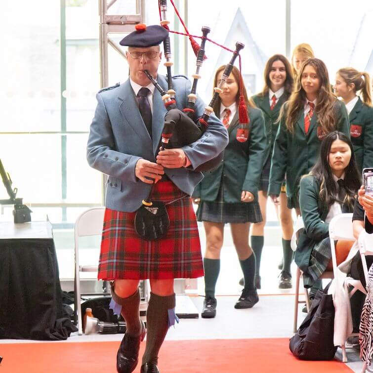 A man in a blue jacket, red tartan kilt, and beret plays bagpipes while walking on a red carpet. Several young women in green blazers and plaid skirts follow behind him indoors.