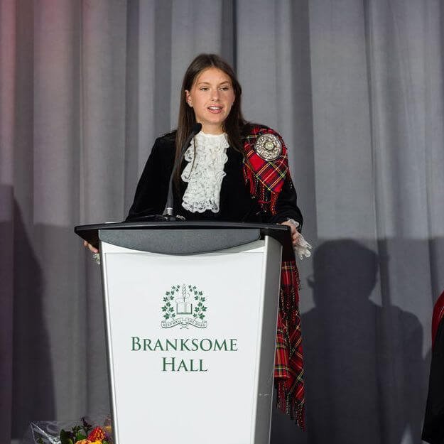 A woman in formal attire with a red tartan sash stands at a podium labeled "Branksome Hall," speaking against a backdrop of gray curtains. Flowers are visible at the base of the podium.