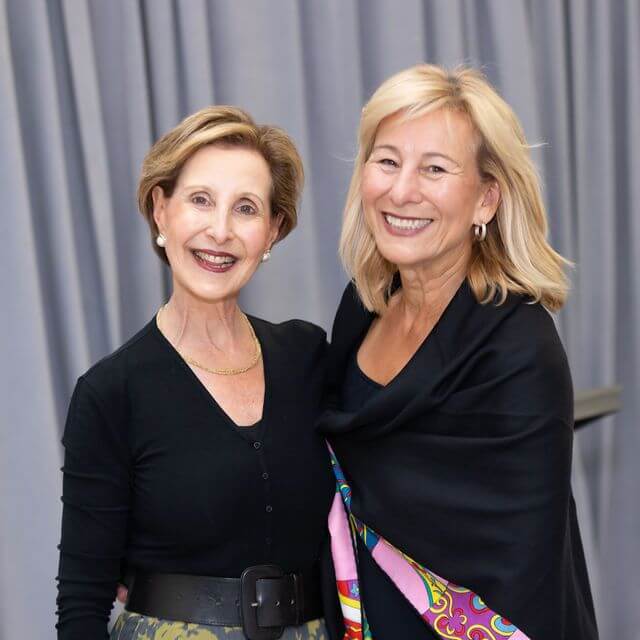 Two smiling women stand close together in front of grey curtains. Both wear black tops; one has a colorful patterned scarf draped over her shoulders. They have light hair and appear happy, posing for the photo.